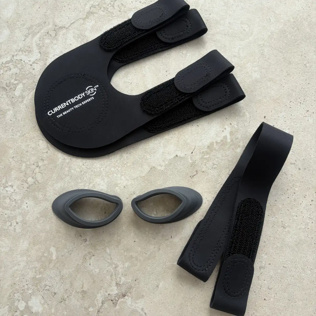 The CurrentBody Series 2 accessories laid out on stone: the branded head strap, the chin strap and the two grey silicone eye inserts
