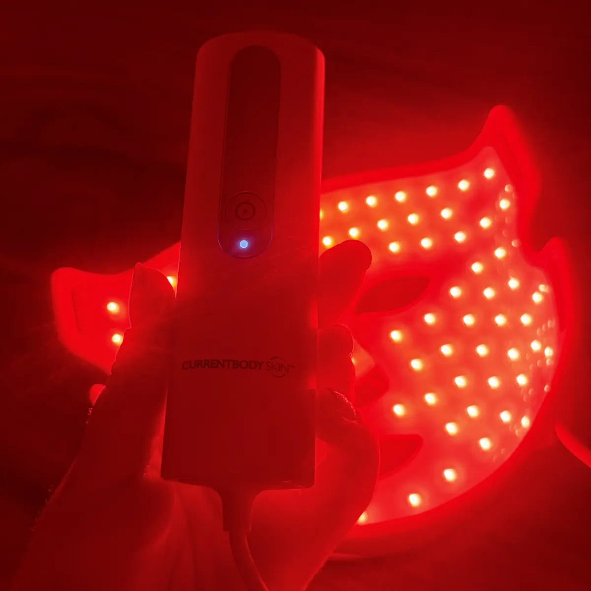 A hand holding a lit CurrentBody controller in front of a LED mask glowing red behind it