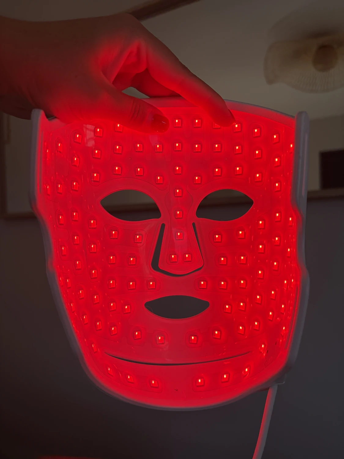 The CurrentBody Series 2 mask lit in red and held up in a dark living room, every diode of the grid visible